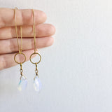 a model holding the model wearing the Aurora Earrings by Third & Co. Studio: faceted synthetic wavy-shaped Opalite in clear and blue flashing rainbow colors, small sections of gold plated vintage circle geometric chain, gold-plated stainless steel ear wires, lightweight sparkly statement earrings, shown against a white background