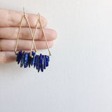 a model holding the Axis Earrings by Third & Co. Studio: Lapis shards in blue and cream, triangular shape raw brass frames, and 14k gold filled ear wires. shown against a white backdrop
