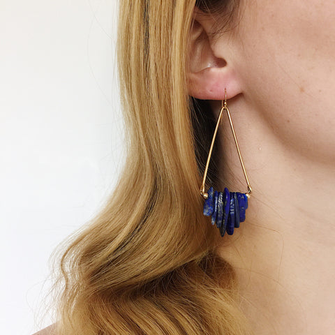 a model wearing the Axis Earrings by Third & Co. Studio: Lapis shards in blue and cream, triangular shape raw brass frames, and 14k gold filled ear wires, shown against a white backdrop