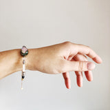 model's wrist wearing a Bracelet with blue green black abalone, white and cream yellow and tan mother of pearl, gray hematite, clear faceted quartz, sterling silver beads and chain adjustable length on gray background