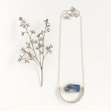 The Ara Necklace by Third & Co. Studio: a single flat irregular-shaped Kyanite nugget in blue gray and black, silver plated crescent shape with oxidized spots, and nickel-free silver plated chain long 32" length, shown against a plain white background and dried florals