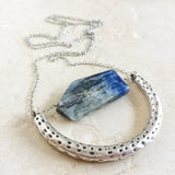 The Ara Necklace by Third & Co. Studio: a single flat irregular-shaped Kyanite nugget in blue gray and black, silver plated crescent shape with oxidized spots, and nickel-free silver plated chain long 32" length, shown against a beige background