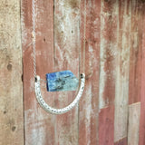a close up shot of the The Ara Necklace by Third & Co. Studio: a single flat irregular-shaped Kyanite nugget in blue gray and black, silver plated crescent shape with oxidized spots, and nickel-free silver plated chain long 32" length, shown against a rustic red background
