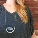 a model in a gray v-neck top with long hair wearing the The Ara Necklace by Third & Co. Studio: a single flat irregular-shaped Kyanite nugget in blue gray and black, silver plated crescent shape with oxidized spots, and nickel-free silver plated chain long 32" length, shown against a rustic red background