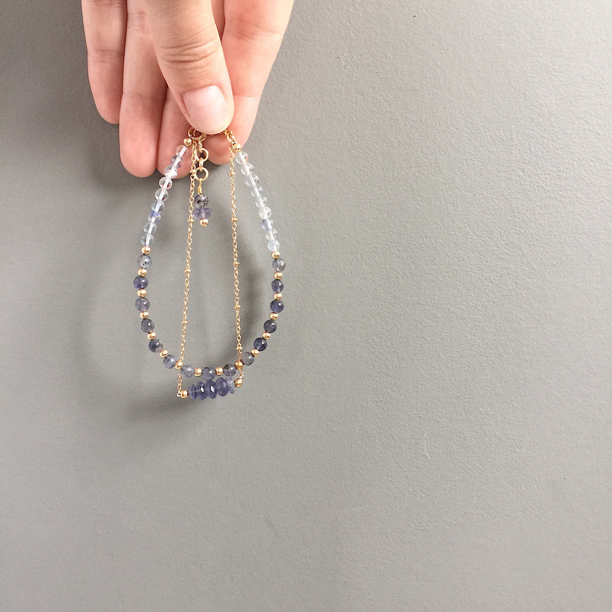 a model holding the Andromeda Bracelet by Third & Co. Studio; faceted Iolite, round Quartz and Iolite, gold plated satellite chain adjustable length bracelet with lobster claw clasp, shown against a gray background