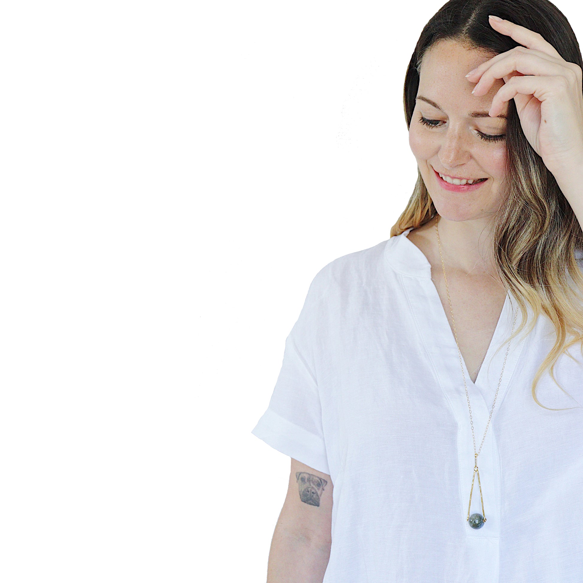 a model in a while n-neck top and long hair wearing a Round Labradorite in green, gray and flashes of blue with an elongated triangular hammered brass frame, a small vintage bar accent and 14K gold fill chain 30" length necklace
