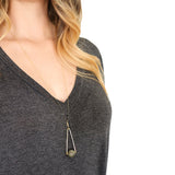 a model in a gray v-neck dress with long hair wearing Round Labradorite in green, gray and flashes of blue with an elongated triangular hammered brass frame, a small vintage bar accent and 14K gold fill chain 30" length necklace
