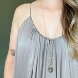 a model in a gray dress with long hair wearing Round Labradorite in green, gray and flashes of blue with an elongated triangular hammered brass frame, a small vintage bar accent and 14K gold fill chain 30" length necklace