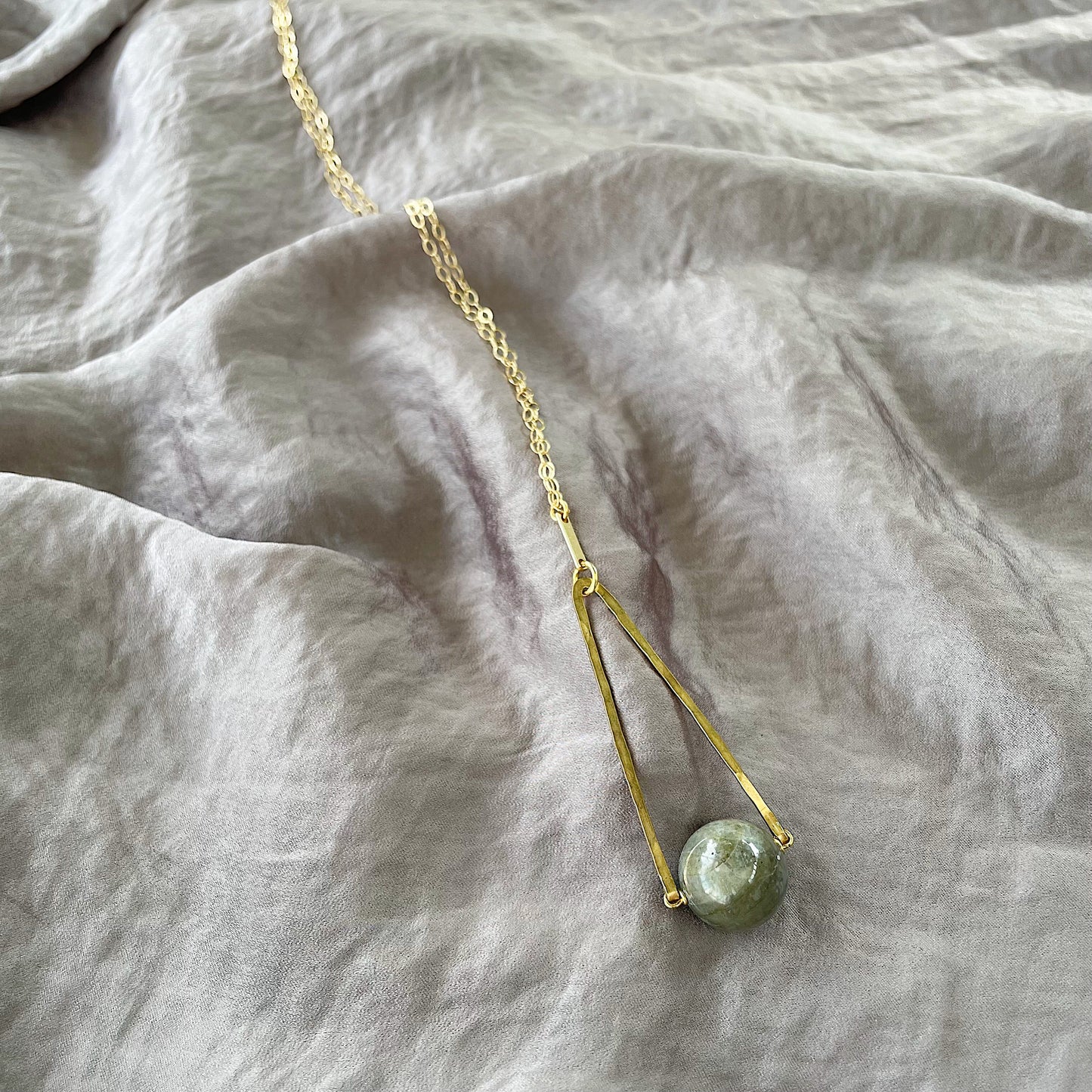 Round Labradorite in green, gray and flashes of blue with an elongated triangular hammered brass frame, a small vintage bar accent and 14K gold fill chain 30" length necklace on a silky gray fabric background