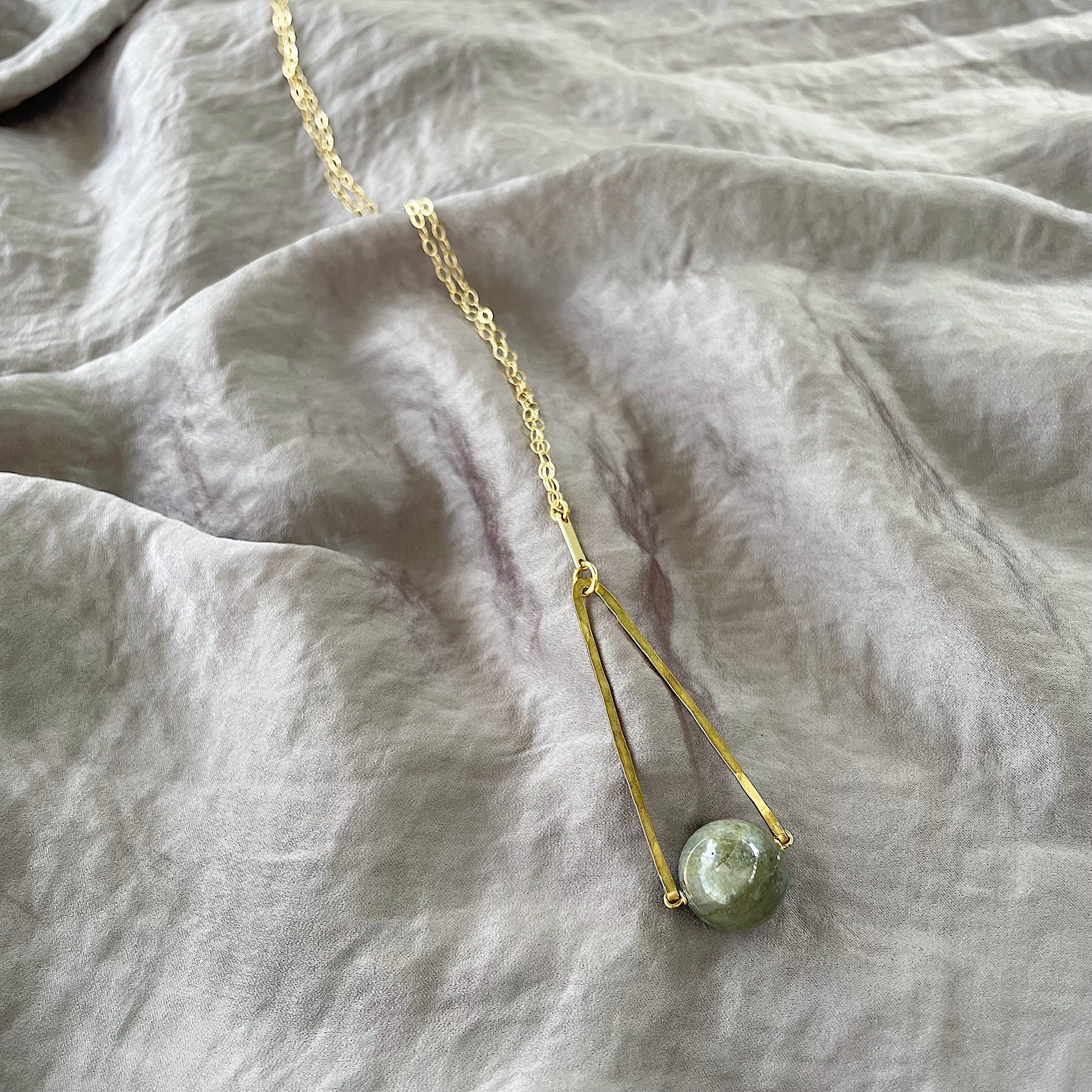 Round Labradorite in green, gray and flashes of blue with an elongated triangular hammered brass frame, a small vintage bar accent and 14K gold fill chain 30" length necklace on a silky gray fabric background