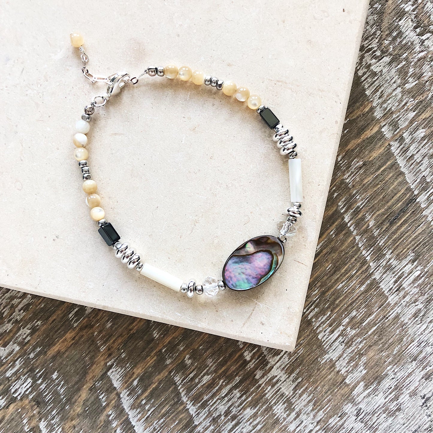 Bracelet with blue green black abalone, white and cream yellow and tan mother of pearl, gray hematite, clear faceted quartz, sterling silver beads and chain adjustable length on textured wood and stone tile background