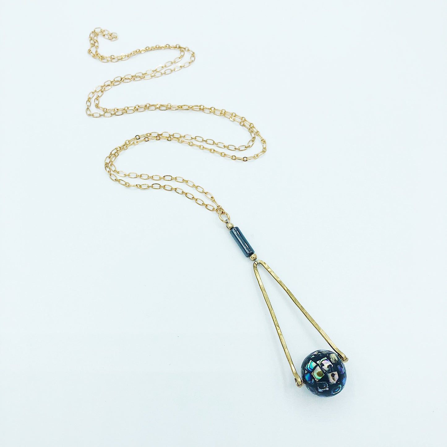 Round Abalone "disco ball" in green, black, cream, tan, brown, and blue with elongated hammered brass triangle frame, gray Hematite round tube, and gold plated nickel-free chain long lnegth 30" chain necklace on white background