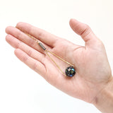 a hand holding a Round Abalone "disco ball" in green, black, cream, tan, brown, and blue with elongated hammered brass triangle frame, gray Hematite round tube, and gold plated nickel-free chain long lnegth 30" chain necklace on white background