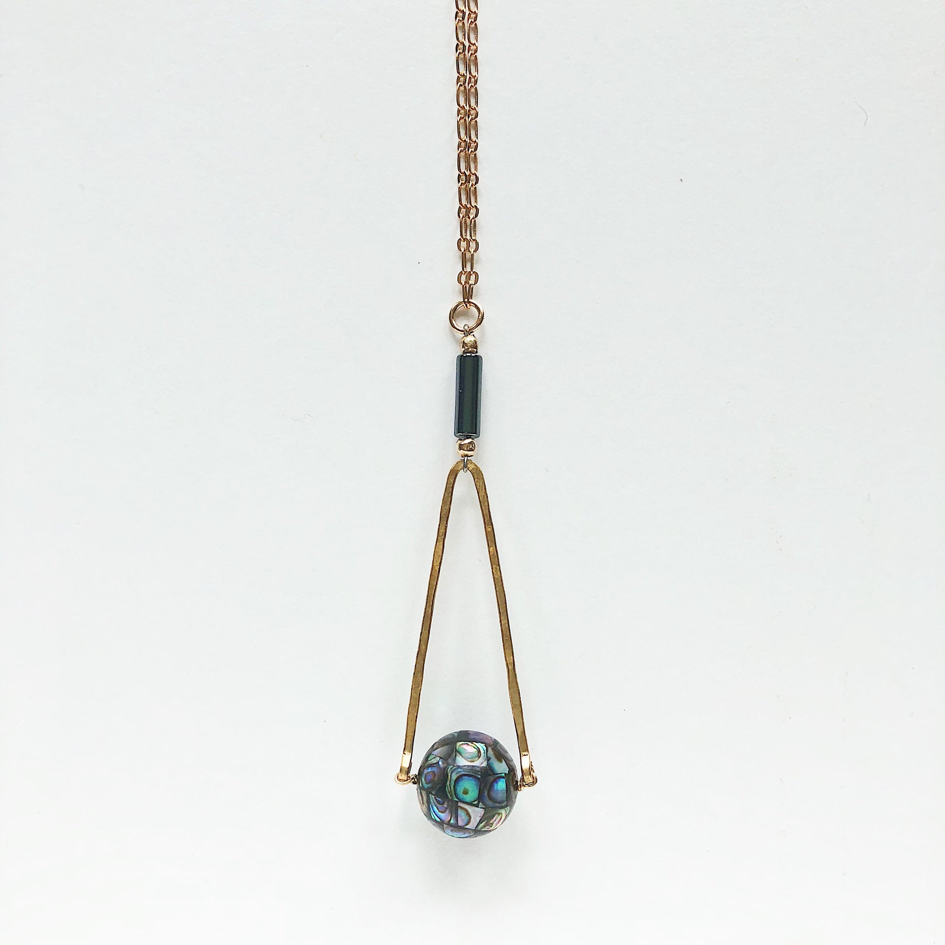 Round Abalone "disco ball" in green, black, cream, tan, brown, and blue with elongated hammered brass triangle frame, gray Hematite round tube, and gold plated nickel-free chain long lnegth 30" chain necklace on white background