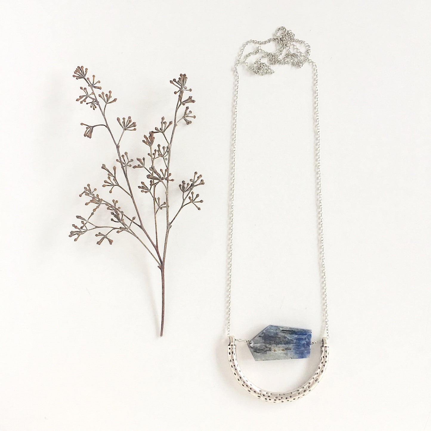 The Ara Necklace by Third & Co. Studio: a single flat irregular-shaped Kyanite nugget in blue gray and black, silver plated crescent shape with oxidized spots, and nickel-free silver plated chain long 32" length, shown against a plain white background and dried florals