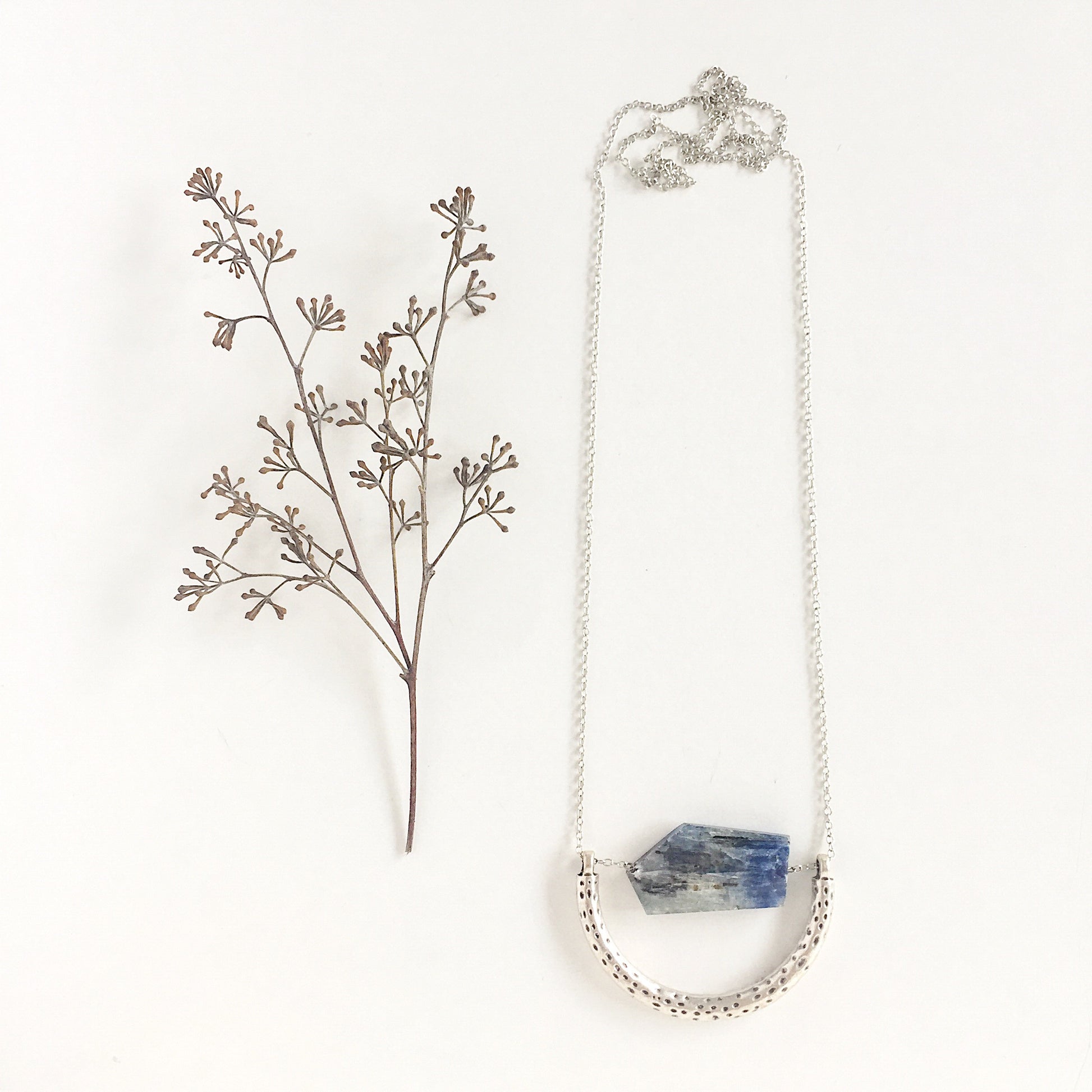 The Ara Necklace by Third & Co. Studio: a single flat irregular-shaped Kyanite nugget in blue gray and black, silver plated crescent shape with oxidized spots, and nickel-free silver plated chain long 32" length, shown against a plain white background and dried florals