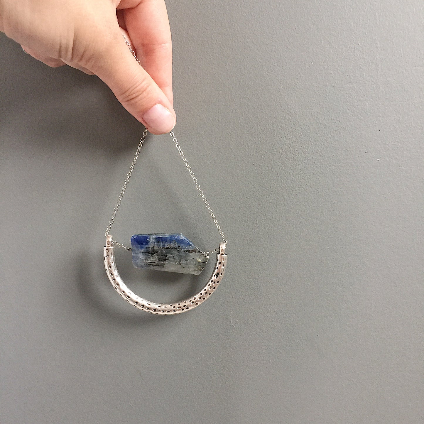 a model holding the The Ara Necklace by Third & Co. Studio: a single flat irregular-shaped Kyanite nugget in blue gray and black, silver plated crescent shape with oxidized spots, and nickel-free silver plated chain long 32" length, shown against a plain gray background