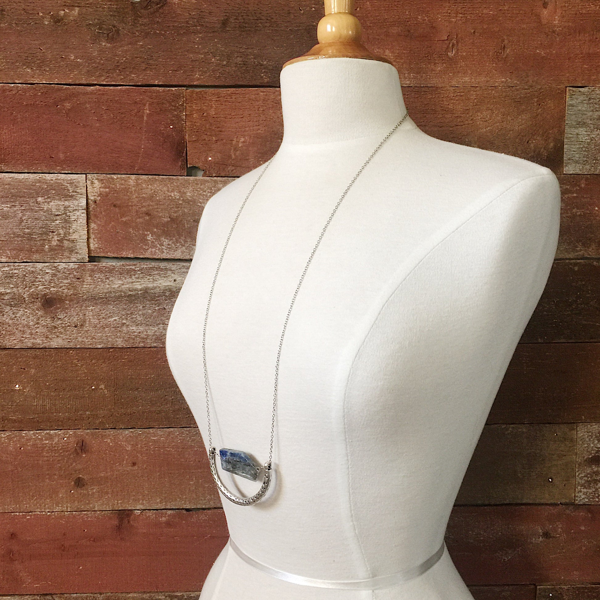 The Ara Necklace by Third & Co. Studio: a single flat irregular-shaped Kyanite nugget in blue gray and black, silver plated crescent shape with oxidized spots, and nickel-free silver plated chain long 32" length, shown on a beige mannequin against a rustic red background
