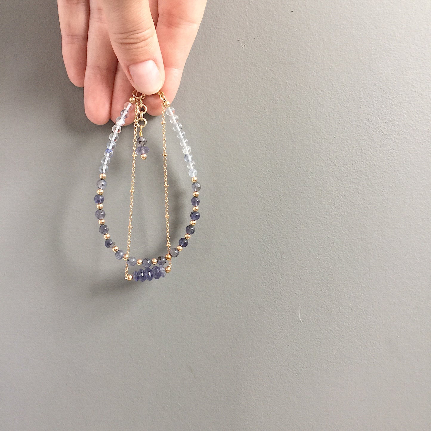 a model holding the Andromeda Bracelet by Third & Co. Studio; faceted Iolite, round Quartz and Iolite, gold plated satellite chain adjustable length bracelet with lobster claw clasp, shown against a gray background