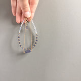 a model holding the Andromeda Bracelet by Third & Co. Studio; faceted Iolite, round Quartz and Iolite, gold plated satellite chain adjustable length bracelet with lobster claw clasp, shown against a gray background