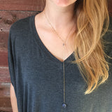 a model in a gray v-neck top and long hair, wearing the Andromeda Necklace by Third & Co. Studio; faceted and round blue Iolite, gold plated satellite chain lariat necklace with lobster claw clasp, shown against a rustic red background