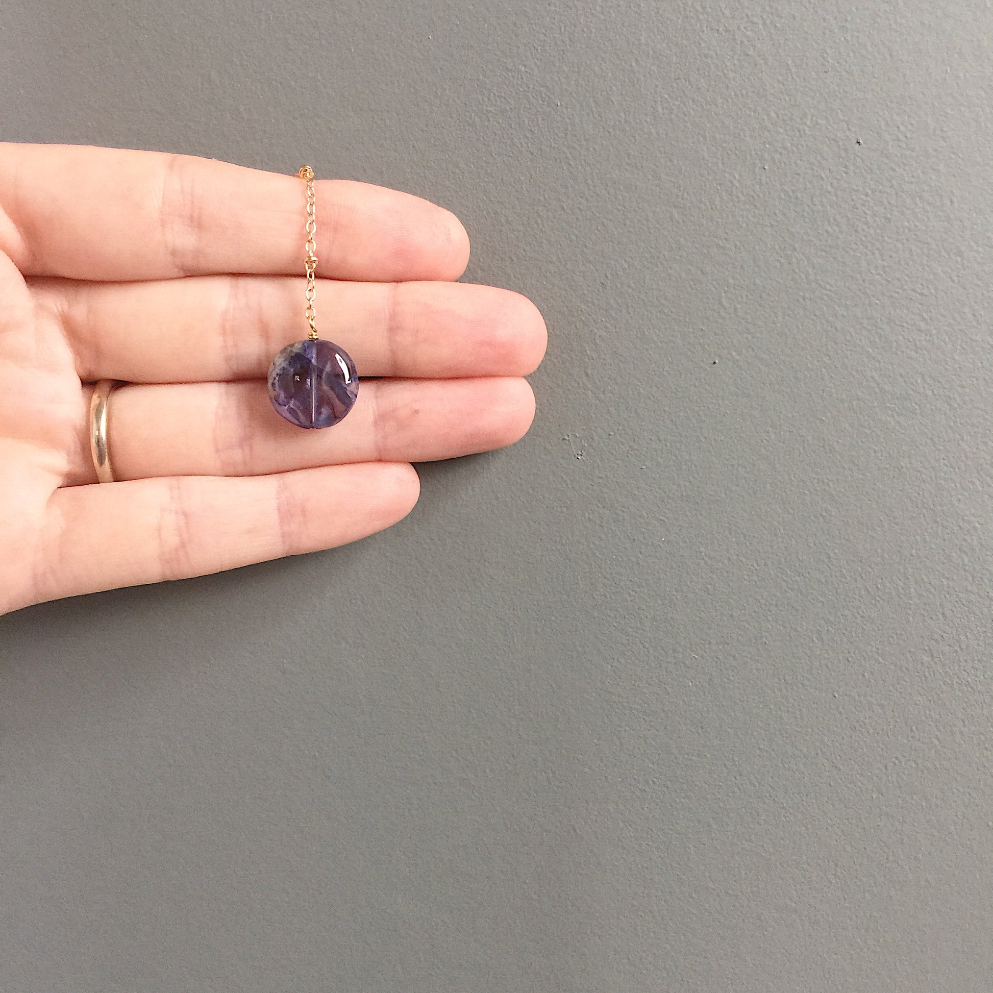 a model holding the lariat of the Andromeda Necklace by Third & Co. Studio; faceted and round blue Iolite, gold plated satellite chain lariat necklace with lobster claw clasp, shown against a gray background