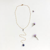 Andromeda Necklace by Third & Co. Studio; faceted and round blue Iolite, gold plated satellite chain lariat necklace with lobster claw clasp, shown against a white background with purple dried flowers