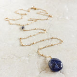 Andromeda Necklace by Third & Co. Studio; faceted and round blue Iolite, gold plated satellite chain lariat necklace with lobster claw clasp, shown against a beige background