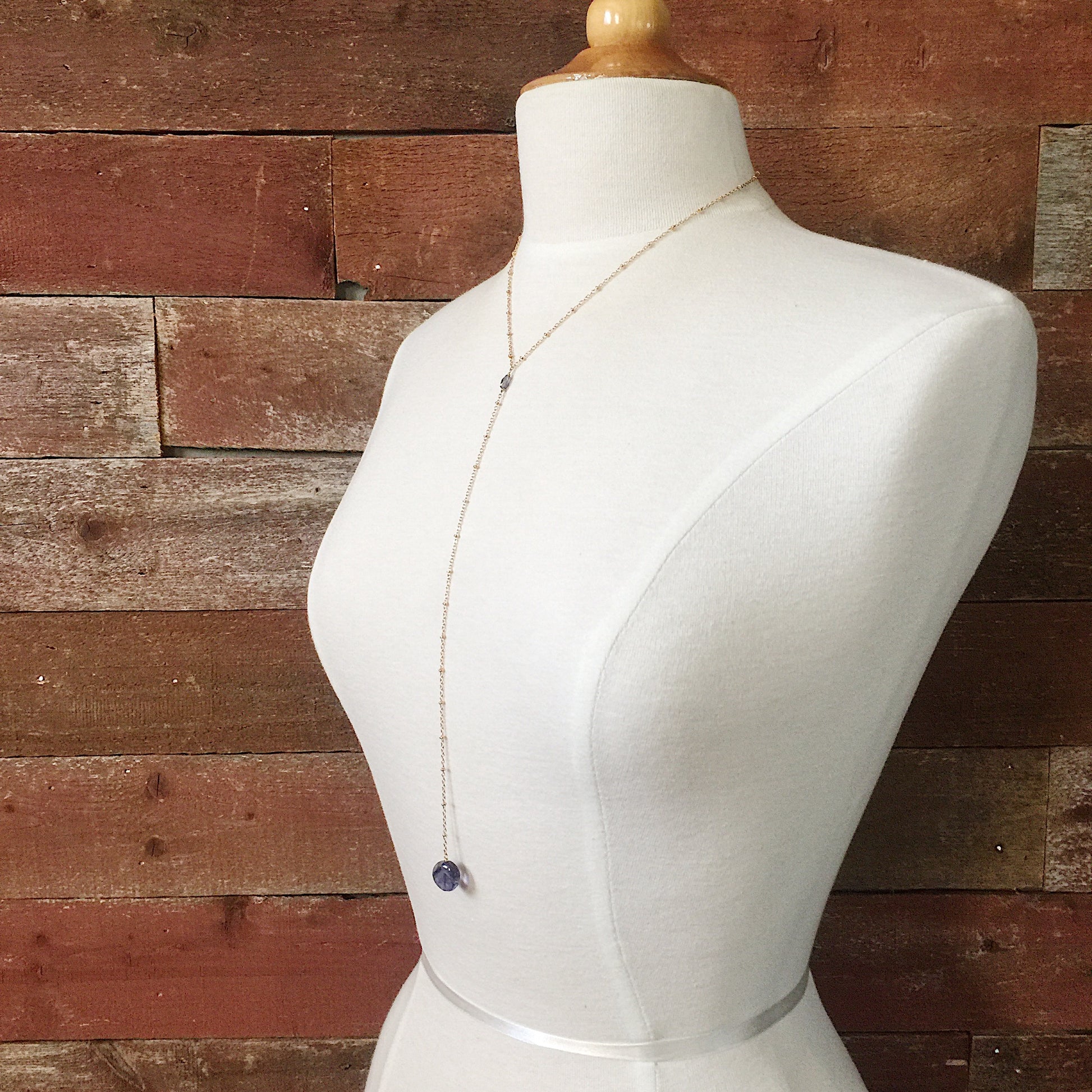 Andromeda Necklace by Third & Co. Studio; faceted and round blue Iolite, gold plated satellite chain lariat necklace with lobster claw clasp, shown on a mannequin against a rustic red background