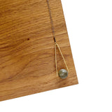 Round Labradorite in green, gray and flashes of blue with an elongated triangular hammered brass frame, a small vintage bar accent and 14K gold fill chain 30" length necklace on a wood and white background