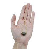 A hand holding a Round Labradorite in green, gray and flashes of blue with an elongated triangular hammered brass frame, a small vintage bar accent and 14K gold fill chain 30" length necklace