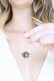 a model holding up a Round Labradorite in green, gray and flashes of blue with an elongated triangular hammered brass frame, a small vintage bar accent and 14K gold fill chain 30" length necklace
