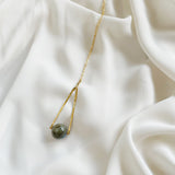 Round Labradorite in green, gray and flashes of blue with an elongated triangular hammered brass frame, a small vintage bar accent and 14K gold fill chain 30" length necklace laying on cream colored silky fabric